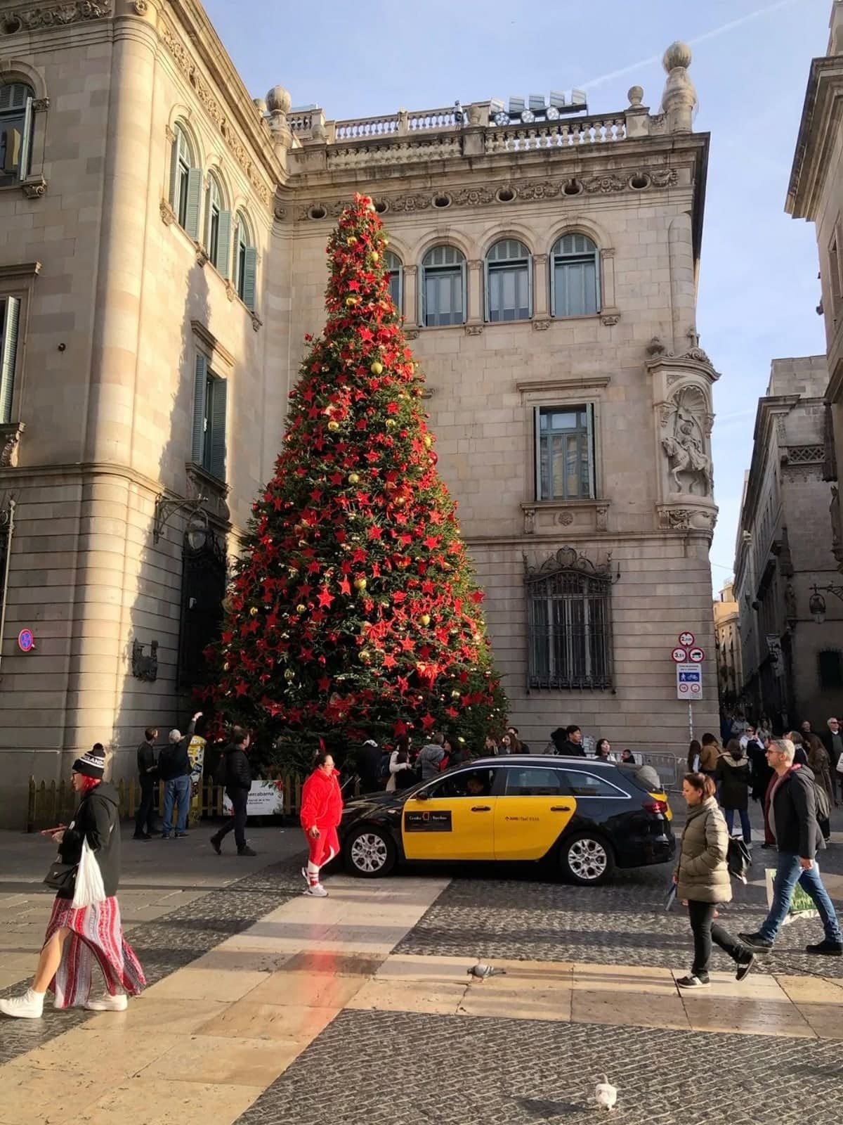 A tall red Christmas tree in a Barcelona square, a yellow and black taxi passing