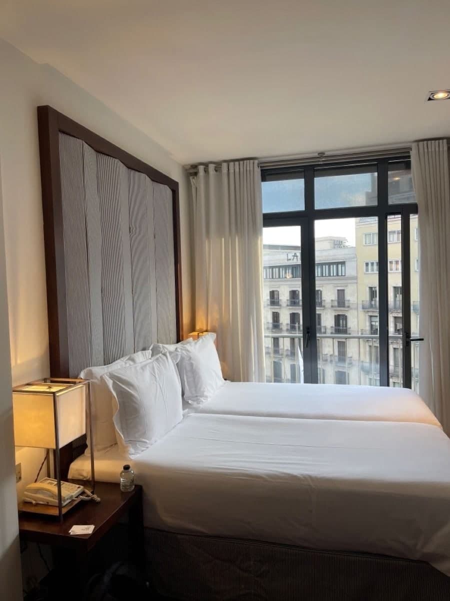 A white hotel bedroom with a large window onto Barcelona facades
