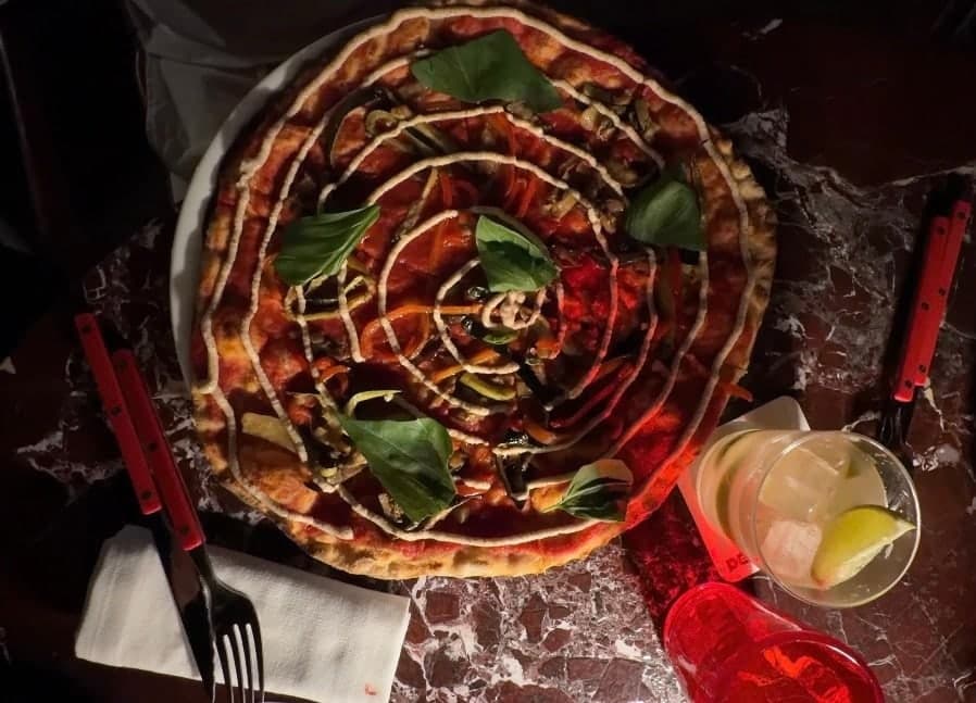 A pizza with basil on a red marble table, a drink beside it, from above