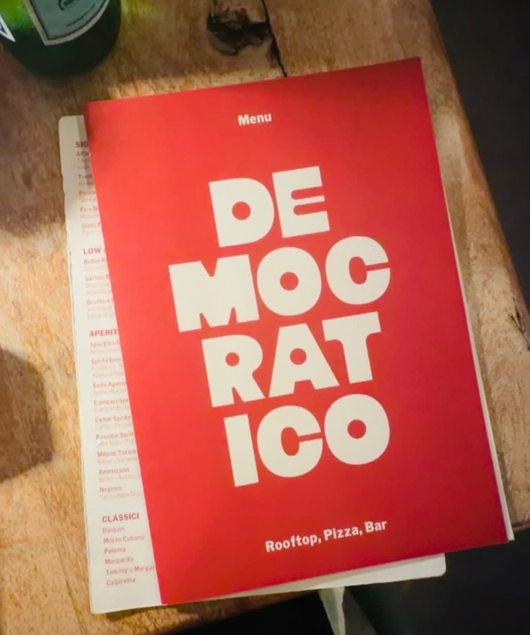 The red Democratico menu on a wooden table, reading Rooftop, Pizza, Bar
