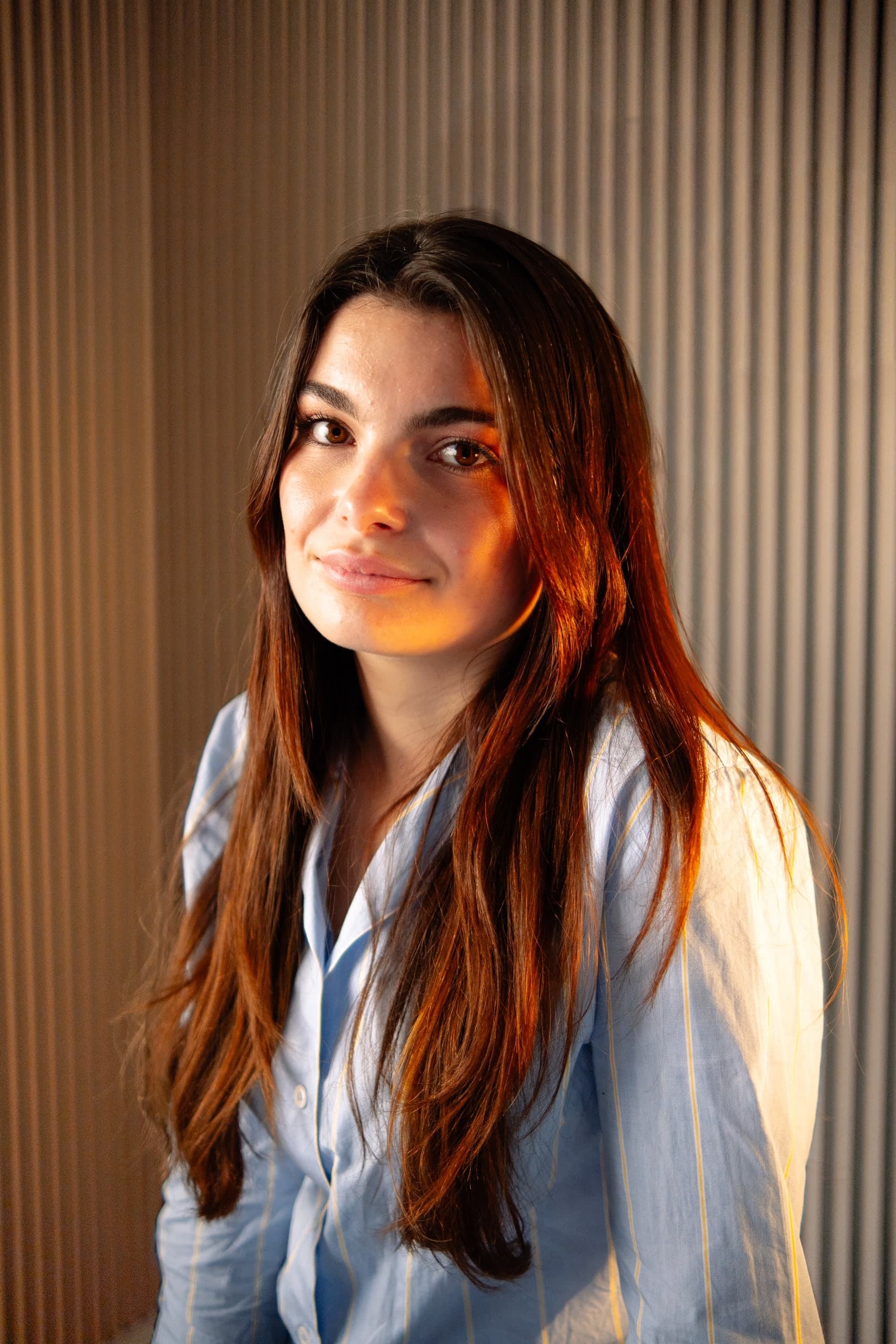 Eleonora Cavinato smiling, in a light blue striped shirt, against a ribbed curtain in warm light