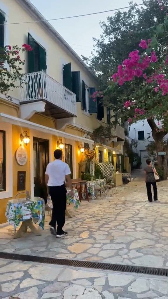 A waiter walking down a stone-paved village street at dusk, past bougainvillea and tables outside the tavernas