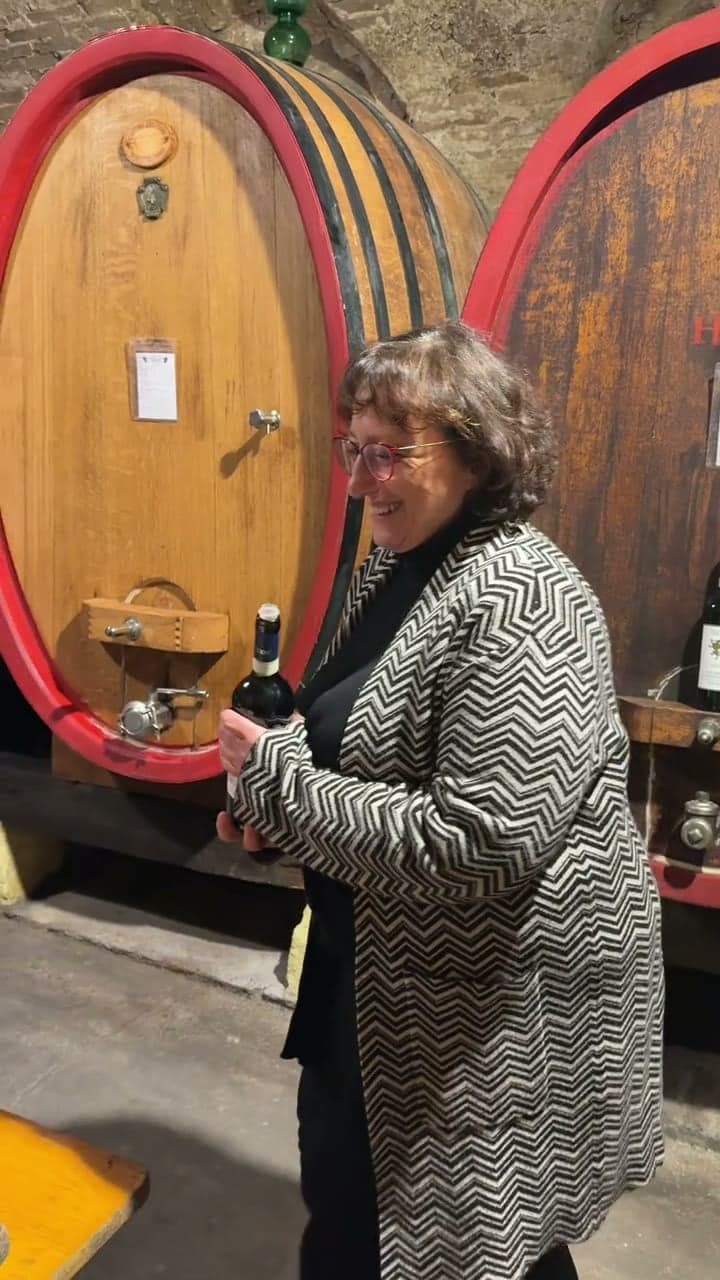 A wine cellar visit: bottles in straw, huge oak barrels with red rims, a long tasting table under the vaults, the host holding a bottle, cured meats and cheese, and red wine poured into a glass